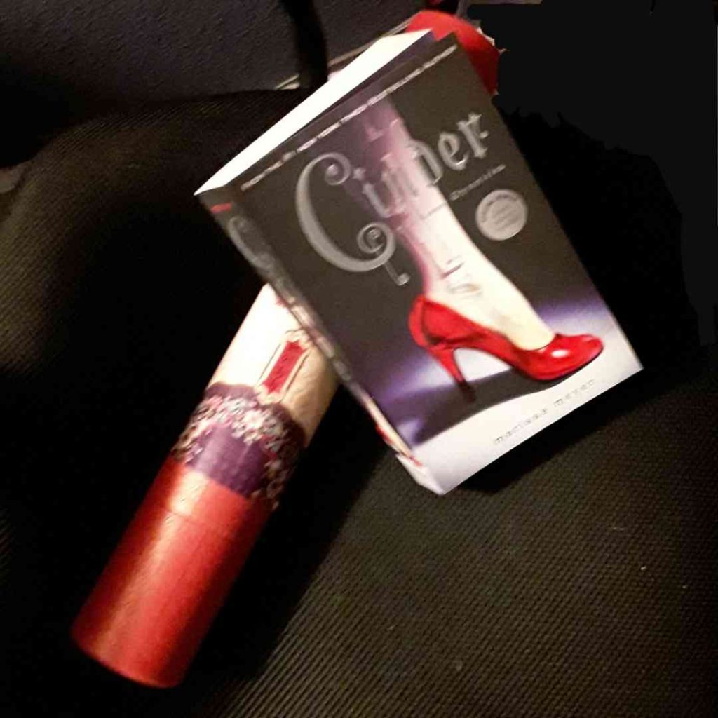 A paperback copy of Cinder rests on a red poster tube. On the cover, a woman's leg in a red pump has mechanical parts showing through the skin.
