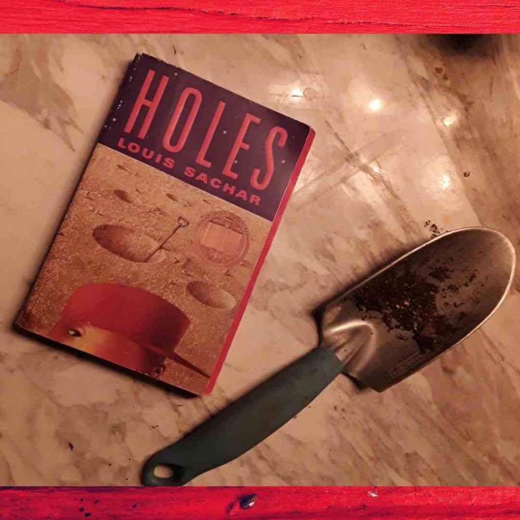 A paperback copy of Holes sits next to a dirty trowel. On the cover, there's a lunar-like landscape the top of a boy's head near the bottom. He's wearing a red cap.